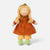 A handmade fabric doll with long blonde braided hair, wearing an orange dress, yellow pants, green crocheted shoes, and a gold bonnet, set against a plain light gray background.