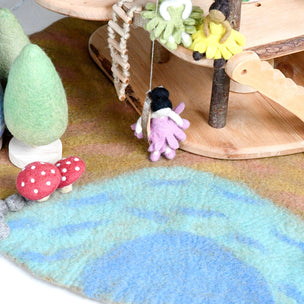 A close-up of a handcrafted felted wool miniature scene featuring a small fairy in a purple and black dress near a wooden platform, with felted trees, red-and-white mushrooms, and a blue-green ground mat.