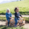 Two children riding wooden toy trucks along a grassy track.
