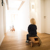 Child riding a wooden toy lorry in a bright room with white walls and wooden floor.