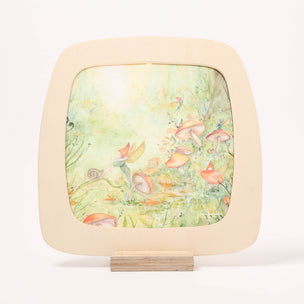 A square framed Toverlux Window frame with rounded corners, featuring a whimsical watercolor forest scene with mushrooms, flowers, and a fairy, displayed on a small wooden stand against a white background.