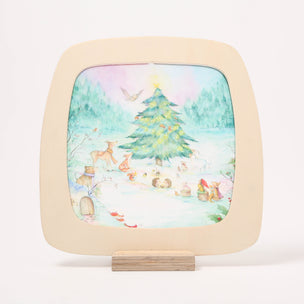 A square wooden toverlux window frame with rounded corners, featuring a hand-painted winter forest scene with a Christmas tree, deer, and small woodland figures. The plaque is displayed on a small wooden stand against a clean white background.