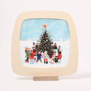 A square decorative Toverlux window frame with rounded corners, displayed on a small wooden stand against a plain white background. The plate features a cream-colored border and a colorful illustration of children gathered around a decorated Christmas tree in the snow.