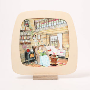 A square decorative toverlux window frame with rounded corners and a light beige border, displayed on a small wooden stand. The center of the plate features a colorful illustration of a cozy cabin living room with a fireplace, sofa, bookshelves, and a balcony.