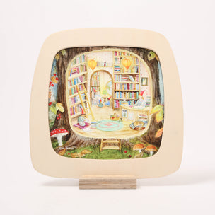 A square decorative Toverlux Window frame with rounded corners, displayed on a small wooden stand against a clean white background. The plate features a cream-colored border and a colorful, whimsical illustration of a cozy book-filled treehouse scene with a ladder, shelves, and small characters.