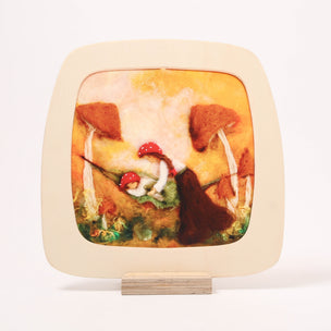 A square wooden toverlux frame rounded corners displaying a handcrafted felt artwork of a fairy among red-capped mushrooms, supported by a small wooden stand against a white background.