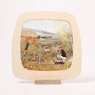 A square decorative Toverlux Frame with rounded corners, displayed on a small wooden stand. The plate features a cream-colored border and a colorful hand-painted illustration of a pastoral landscape with children, sheep, and trees.