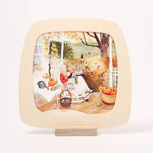A toverlux frame with rounded corners, displayed on a small wooden stand against a clean white background. The plate features a colorful illustration of a cozy autumn porch scene with a woman in a rocking chair, pumpkins, baskets, and children playing outdoors.