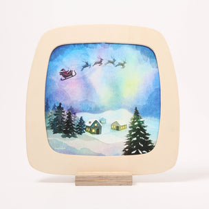 A square toverlux frame with rounded corners, mounted on a small wooden stand. The plaque has a light natural wood frame and features a colorful winter scene printed in the center, showing Santa Claus in a sleigh with reindeer flying over snow-covered houses and evergreen trees under a blue and purple sky.