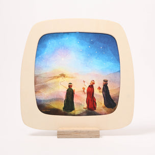 A square wooden toverlux frame with rounded corners, featuring a painted scene of three wise men under a starry night sky, displayed on a small wooden stand against a white background.