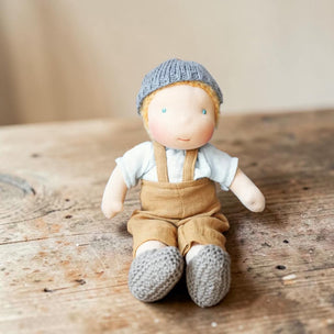 A handmade soft fabric doll with a knitted blue-gray hat, white shirt, tan overalls, and matching knitted booties, sitting on a rustic wooden surface.
