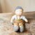 A handmade soft fabric doll with a knitted blue-gray hat, white shirt, tan overalls, and matching knitted booties, sitting on a rustic wooden surface.