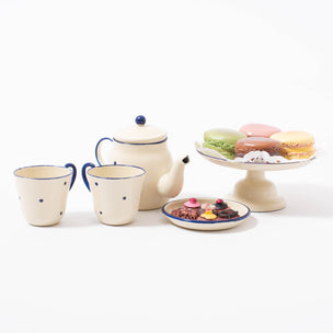 Four different coloured/flavoured resin macarons from Maileg on a hand painted cream metal cake stand with blue detailing and decorative paper napkin, alongside matching teapot and mugs and plate of little cakes | © Conscious Craft